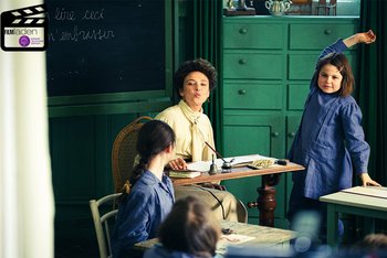 Maria Montessori sitzt an einem Lehrerpult in einem Klassenzimmer vor einer Tafel und lächelt, einen Kussmund formend, andere Schüler:innen an. Neben ihr zeigt eine Schülerin auf.