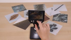 Ein Digitales Mikroskop steht auf einem Tisch. Daneben liegen Bildkarten, auf denen verschiedene Naturmaterialien zu erkennen sind. Eine Hand tippt auf das Digitale Mikroskop.