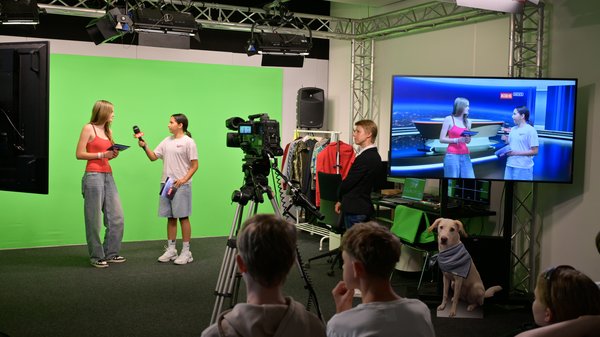 Zwei Schülerinnen stehen mit Moderationskärtchen und Mikrofon in einem TV-Studio vor einem Greenscreen. Eine Erwachsene steht daneben. Schüler:innen sitzen auf Sesseln und schauen zu.