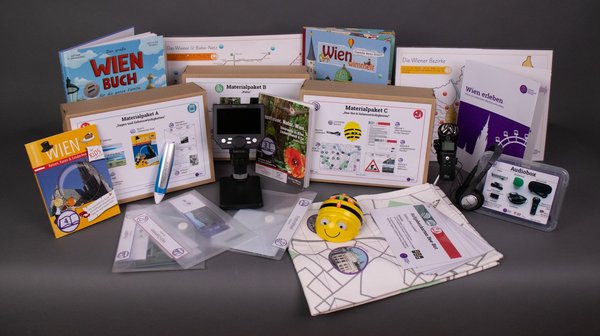 Der Inhalt der Wien erleben-Digibox: Diverse Wien-Bücher für Kinder und pädagogische Materialpakete sowie technische Geräte sind abgebildet. Darunter ein Tellimero-Audiostift, ein Bee-Bot-Roboter, ein Audioaufnahmegerät und ein digitales Mikroskop.