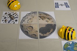 Zwei Lernroboter Bee-Bot auf einer durchsichtigen Matte mit Mond-Motiv und dem Planeten Erde eingelegt