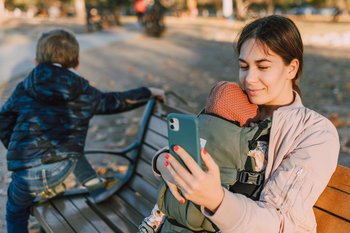 Eine Frau mit Baby in einem Tragegurt sitzt bei herbstlicher Stimmung auf einer Bank und hält ein Smartphone vor sich.