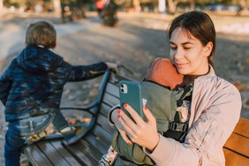 Eine Frau mit Baby in einem Tragegurt sitzt bei herbstlicher Stimmung auf einer Bank und hält ein Smartphone vor sich.