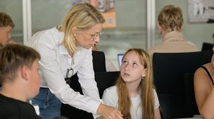 Schüler:innen sitzen an einem Tisch, eine erwachsene Frau beugt sich zu ihnen hinunter und erklärt etwas.